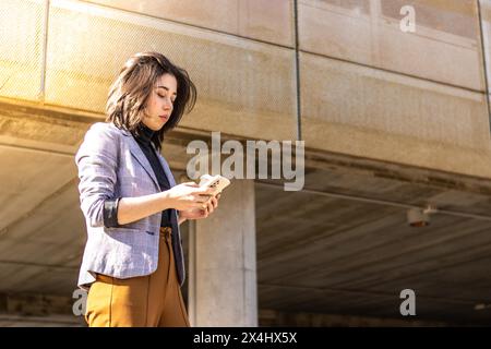 Giovane donna d'affari che cammina per la strada tenendo il cellulare e un bicchiere di caffè, Una donna che cammina lungo la strada tenendo un caffè di vetro e usandolo Foto Stock
