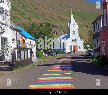 Piccola chiesa e case fiancheggiano una strada decorata con colori arcobaleno, Seydisfjoerdur. Islanda Foto Stock