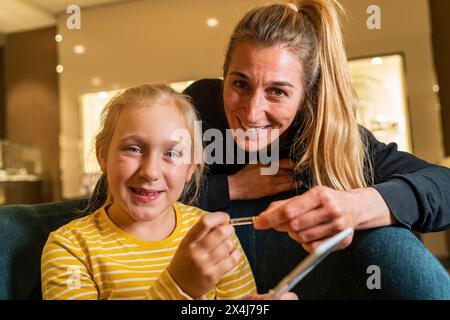 Madre e figlia gioiose che guardano la macchina fotografica, ragazza che tiene nuovi orecchini su un piedistallo e sorride in gioielleria Foto Stock