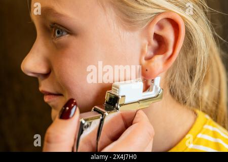 Vista laterale di una ragazza che ottiene il suo orecchio perforato con una pistola perforante da un operatore medico professionista in un negozio di gioielli Foto Stock