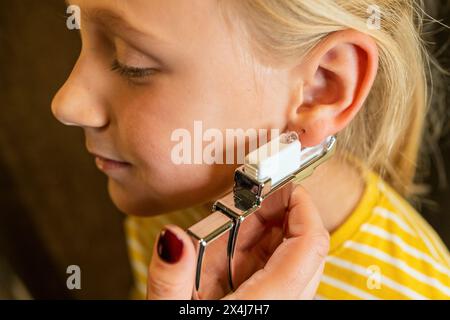 una ragazza che si fa forare l'orecchio con una pistola perforante e unghie rosse visibili, gli occhi chiusi in una gioielleria Foto Stock