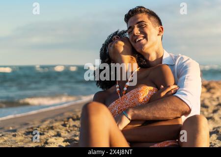 Giovane coppia interrazziale - abbracciando amorevolmente la spiaggia, condividendo un momento di gioia sullo sfondo delle onde dell'oceano al tramonto - romantico e intimo. Foto Stock