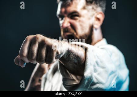 Lottatore di karate che pratica il pugno in palestra. Focalizzazione selettiva sul pugno. Forza e motivazione. Uomo urlante in kimono pronto per l'allenamento di karate. Arrabbiato Foto Stock