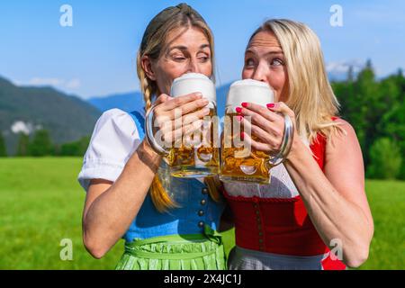 Due donne nel Tracht tradizionale, Dirndl bevono birra frizzante dalle tazze con montagne alpine sullo sfondo, concetto Oktoberfest Foto Stock