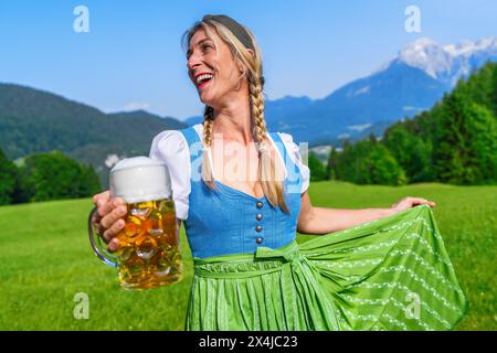 Donna in un tradizionale tracht bavarese blu e verde, abito Dirndl con in mano una tazza di birra con montagne bavaresi sullo sfondo, Oktoberfest concept im Foto Stock