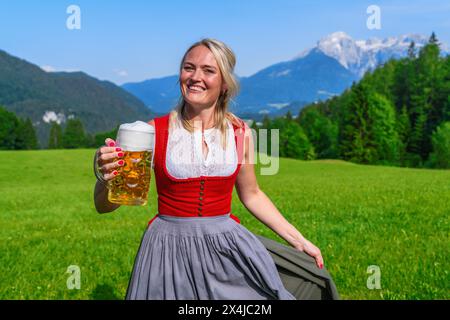 Donna bionda in rosso tradizionale Tracht, Dirndl con una tazza di birra spumante con montagne alpine sullo sfondo, immagine concettuale dell'Oktoberfest Foto Stock