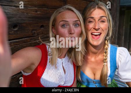 Due donne che scattano selfie di fronte a una cabina alpina in legno sulle montagne bavaresi indossando i tradizionali dirndls o tracht. Oktoberfest bavarese tedesco Foto Stock