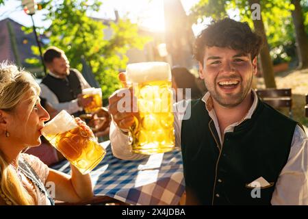 Gioioso giovane che brinda con tazza di birra all'Oktoberfest o all'orto della birra, vestito con abiti tradizionali o tracht in tedesco, con luce solare intensa nel backgroun Foto Stock