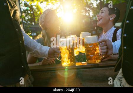 Amici nella tradizionale tostatura tedesca del tracht con tazze bavaresi al tramonto, espressioni felici, in un ambiente all'aperto nel giardino della birra o nell'oktoberfest Foto Stock