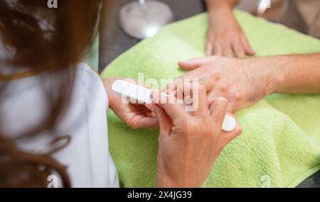 Primo piano di mani maschili durante una manicure, con un manicurista che usa un tampone sulle unghie di un cliente. Foto Stock