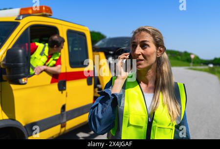 Una donna automobilista parla al telefono dopo che il servizio di traino ha caricato la sua auto su un camion di traino e spiega il suo problema. Concetto di assistenza stradale Foto Stock