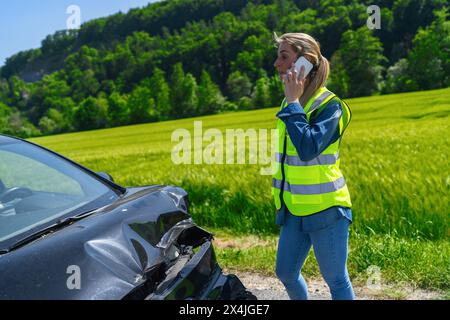 Donna con giubbotto ad alta visibilità al telefono con auto danneggiata in primo piano che mostra il luogo dell'incidente su strada rurale Foto Stock