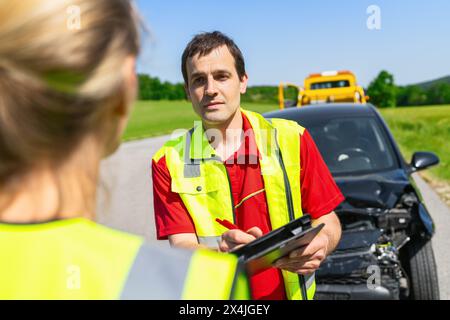 Il lavoratore dell'assistenza stradale che tiene in mano gli appunti discutendo con la donna nel giubbotto di sicurezza dopo un incidente d'auto sul corso successivo Foto Stock