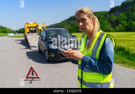 Donna preoccupata o spaventata in un giubbotto di sicurezza con il telefono sul luogo dell'incidente, traino di un camion con la sua auto e triangolo segnaletico sullo sfondo Foto Stock