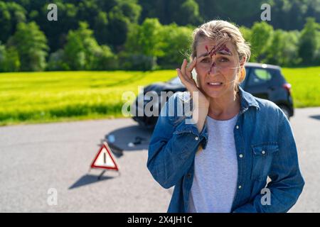 Donna ferita con ferita alla testa in piedi a destra con triangolo di avvertimento e auto schiantata sullo sfondo. Immagine del concetto di assistenza stradale Foto Stock