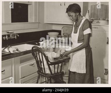 Donna afroamericana in uniforme che lavora come un bambino bianco che nutre domestico seduto su un seggiolone in cucina, Atlanta, Georgia, maggio 1939. Fotografia storica d'epoca della collezione US Government Security Administration anni '1930 Foto: Marion Post Wolcott. Foto Stock