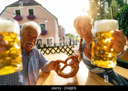 Uomo anziano e giovane donna del tracht bavarese che tiene tazze e pretzel da birra, sorride alla macchina fotografica in una birreria all'aperto o all'Oktoberfest di Monaco, Germania Foto Stock