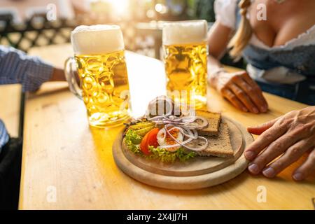 Obatzda tradizionale bavarese con pretzel e ravanelli e tazze da birra, uomo e giovane donna a tracht sullo sfondo presso la birreria all'aperto o l'oktoberfest, Foto Stock