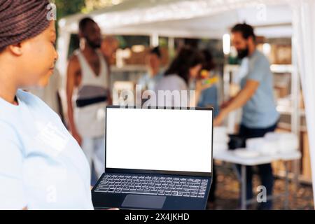 Immagine dettagliata che mostra una donna afroamericana volontaria che trasporta un laptop con un display isolato per la personalizzazione. Donna nera che tiene in mano un minicomputer con un modello di mockup a schermo bianco. Foto Stock