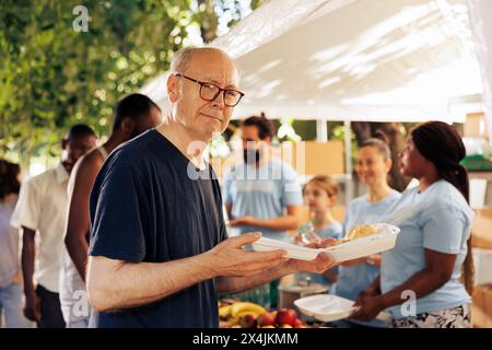 Immagine che mostra un vecchio senzatetto caucasico che tiene il suo pasto dall'organizzazione caritatevole. Gruppo di volontari in una banca alimentare all'aperto che aiuta e alimenta i poveri affamati e i meno privilegiati. Foto Stock