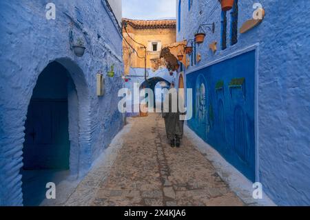 Uomo che cammina su una strada stretta nella medina della città blu. Uomo che indossa abiti tradizionali (Djellaba marocchina), una delle forme più popolari in Marocco. Foto Stock