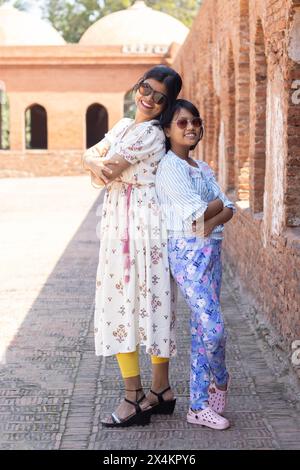 Madre e figlia indiane che indossano gli occhiali da sole, mettono in posa la macchina fotografica dietro le quinte, vicino a un'antica parete di mattoni Foto Stock