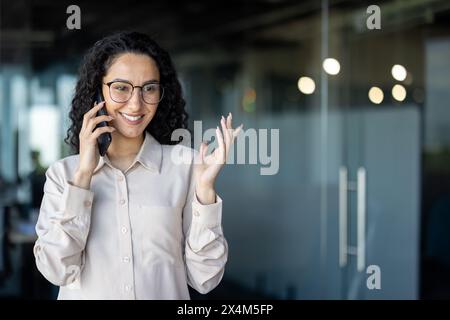 Donna ispanica felice di successo che parla al telefono, donna d'affari sul posto di lavoro all'interno dell'ufficio, lavoratrice con capelli ricci e occhiali, capo donna sicura di sé. Foto Stock