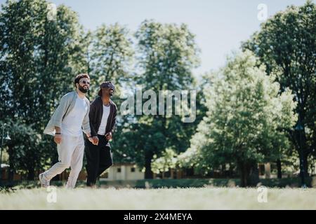Giovane coppia gay multirazziale che si gode una giornata di sole al parco, tenendosi per mano e sorridendo Foto Stock