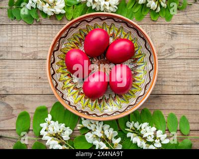 uova di pasqua rosse in un piatto di argilla decorato incorniciato da fiori e foglie di acacia Foto Stock