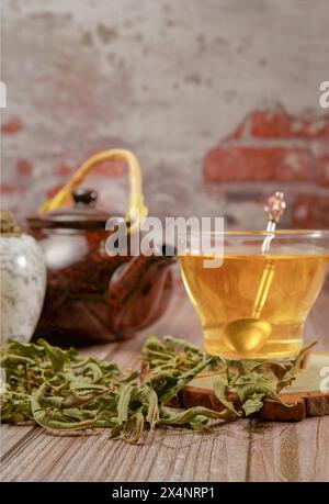 Infusione di verbena di limone in una tazza di vetro con mortaio di marmo e pestello e teiera in ceramica con foglie di verbena di limone essiccate su un tavolo di legno con un Foto Stock