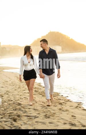 Foto verticale di una giovane coppia caucasica chic che cammina a piedi scalzi tenendosi per mano lungo una spiaggia durante il tramonto Foto Stock