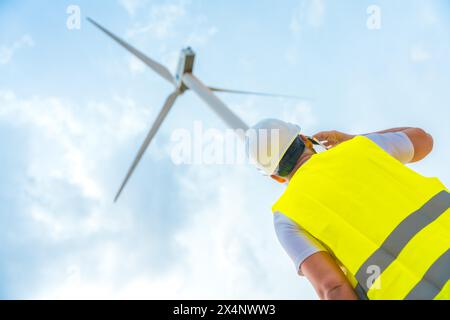 Foto con vista ad angolo basso di un ingegnere caucasico adulto maschio che utilizza il telefono accanto a una turbina eolica in un parco energetico verde Foto Stock