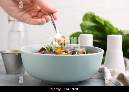 Donna che versa una gustosa salsa a base di aceto (Vinaigrette) dal cucchiaio alla ciotola con insalata al tavolo grigio, primo piano Foto Stock