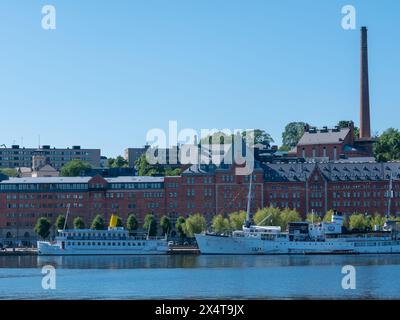 Stoccolma, Svezia - giugno 29 2021: Fiume con traghetti passeggeri bianchi ormeggiati al molo. Grandi edifici industriali in mattoni rossi sullo sfondo. Persone visibili Foto Stock