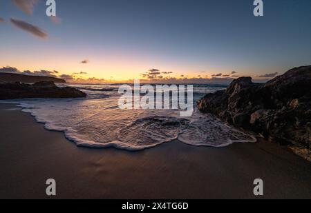 Vista del tramonto sulla spiaggia con le onde che si infrangono delicatamente contro le rocce sotto un cielo colorato Foto Stock