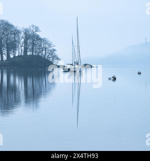 Splendida immagine di paesaggio tranquillo della mattina nebbiosa di primavera su Windermere nel Lake District con barche ormeggiate sul lago e lontane cime nebbiose Foto Stock