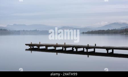 Splendida immagine di paesaggio tranquillo della mattina nebbiosa di primavera su Windermere nel Lake District con barche ormeggiate sul lago e lontane cime nebbiose Foto Stock