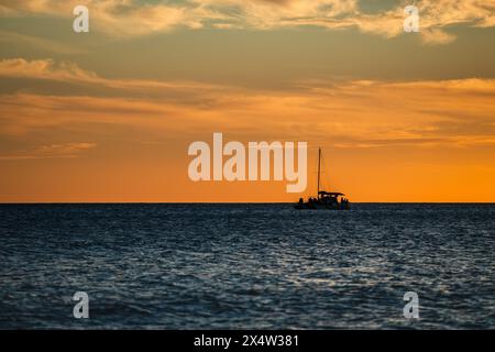 Una barca sta navigando nell'oceano al tramonto. Il cielo è arancione e l'acqua è calma Foto Stock