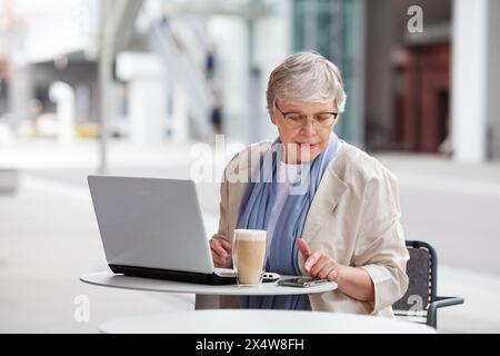 Anziana donna d'affari anziana con capelli corti grigi, lavoratrice freelance matura lavora su un computer portatile, guarda il computer al tavolo in un ritratto all'aperto. Età Foto Stock