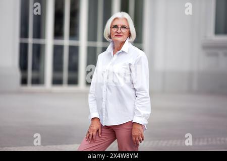 Ritratto di una bella anziana donna con i capelli grigi che indossa occhiali, vestita con una camicia bianca all'aperto in città su uno sfondo luminoso Foto Stock