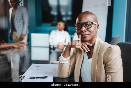 Una donna d'affari africana che indossa gli occhiali conduce con sicurezza una riunione in una sala riunioni con i suoi colleghi mentre discutono documenti e relazioni. Foto Stock