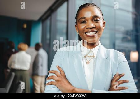 Una giovane donna sicura di sé si trova in una sala riunioni, circondata da uomini d'affari. Ha le braccia incrociate e un sorriso professionale sul viso. Foto Stock
