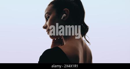 Una giovane donna con auricolari wireless si trova in uno studio e ascolta tranquillamente la sua playlist. La sua espressione rilassata mostra tranquillità. Foto Stock
