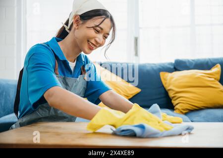 Una giovane donna felice che indossa guanti gialli pulisce e pulisce il tavolo nel suo soggiorno. Impegnato in Foto Stock