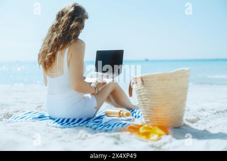 Vista da dietro una donna moderna sulla costa dell'oceano con borsa di paglia, schermo vuoto per laptop e asciugamano a righe. Foto Stock