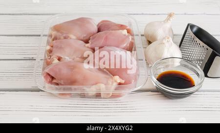 Cosce di pollo crude in un contenitore di plastica, salsa di soia e pressa per aglio e aglio freschi da vicino su uno sfondo di legno bianco Foto Stock