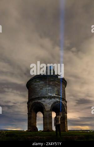 La figura solitaria si trova davanti a un antico mulino a vento, che getta un fascio di luce nel cielo notturno Foto Stock