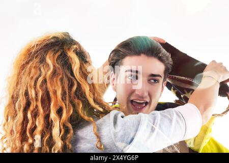 la giovane donna con una giacca gialla ride di cuore mentre regola un cappello festoso sulla testa del suo amico maschio Foto Stock