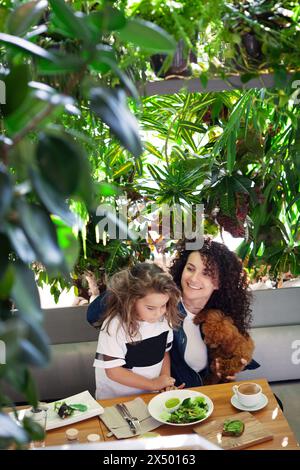 Mamma, figlia e cane, cena in un vivace ristorante vegano, gustando verdure, circondate da una vegetazione lussureggiante e abbracciando lo stile di vita vegetale Foto Stock