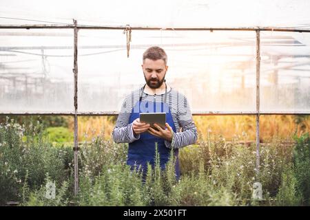 Piccola impresa di serra. Giardiniere, tenendo un tablet che guarda l'ordine, prepara fiori e piantine per il cliente. Foto Stock